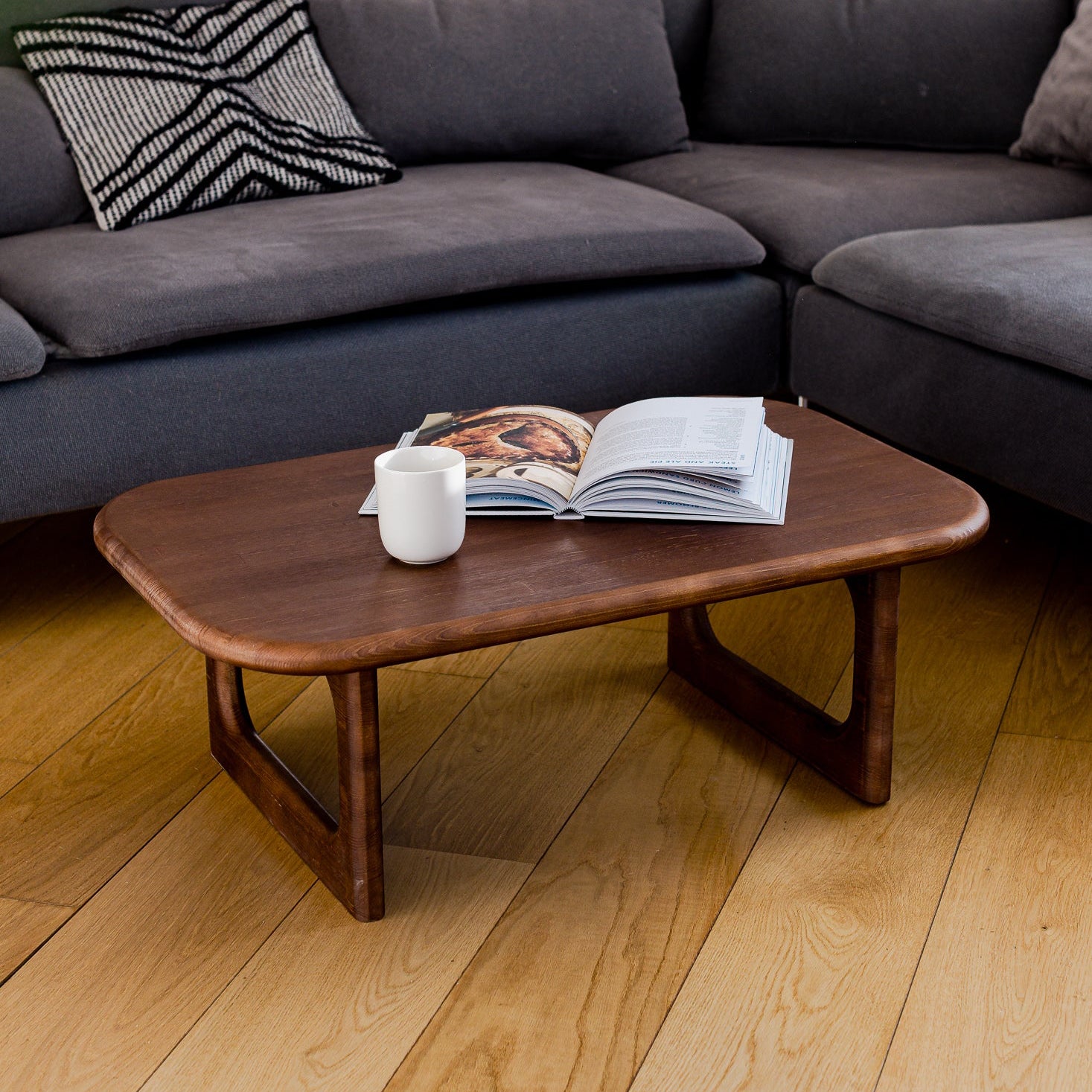 Gray sectional sofa with a wooden coffee table and magazines on a wooden floor.